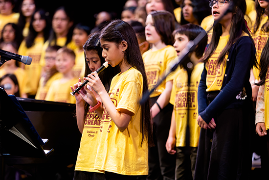 A student with long dark hair plays recorder in front of her choir. All students are wearing yellow t-shirts with 